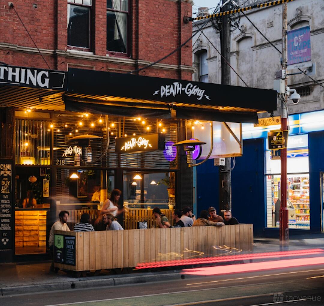 A street-facing bar with outdoor seating, hanging string lights, and wood panel fencing at Death or Glory.