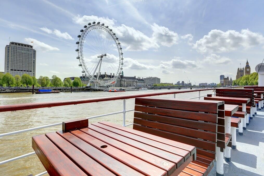 An open boat deck with wooden benches and tables overlooking the Thames and the London Eye at The Tattershall Castle.