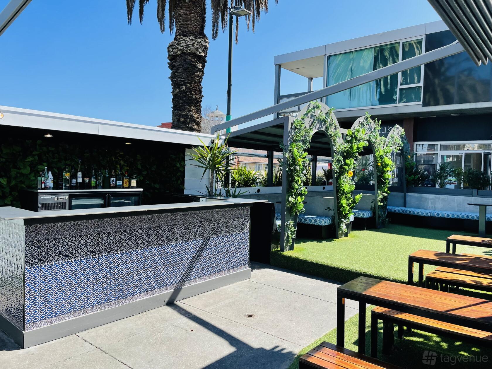 An outdoor bar with patterned tile counter, greenery, covered seating, and picnic tables at Secret Garden Bar.