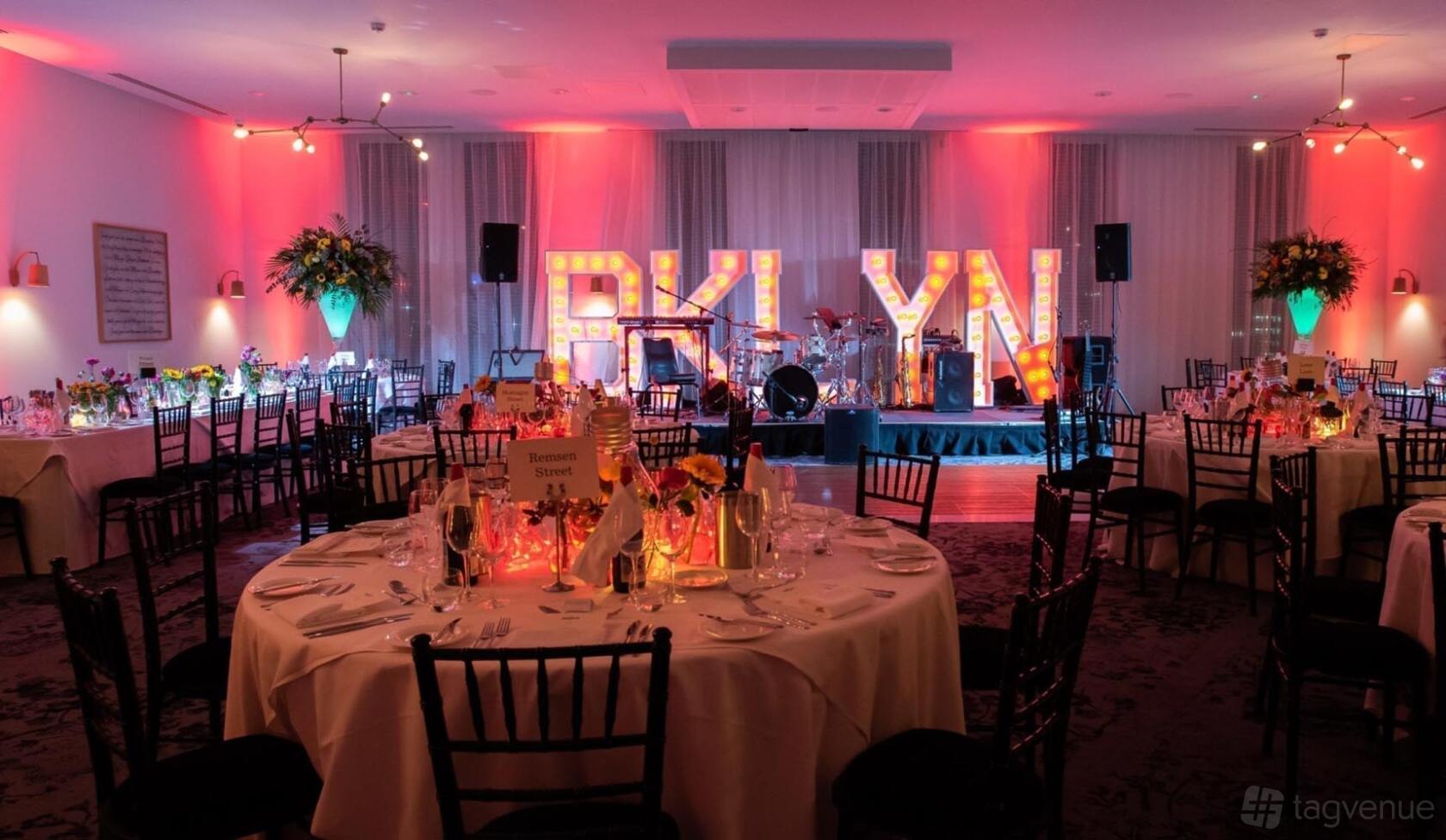 A hotel hall with round tables, black chairs, stage lighting, and illuminated marquee letters at voco Manchester.