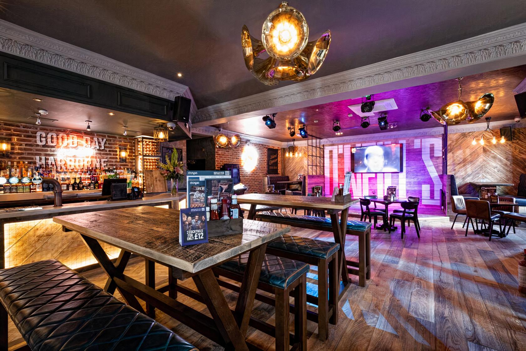 A pub with exposed brick walls, wooden tables, bench seating, and illuminated signage at O'Neill's Harborne.