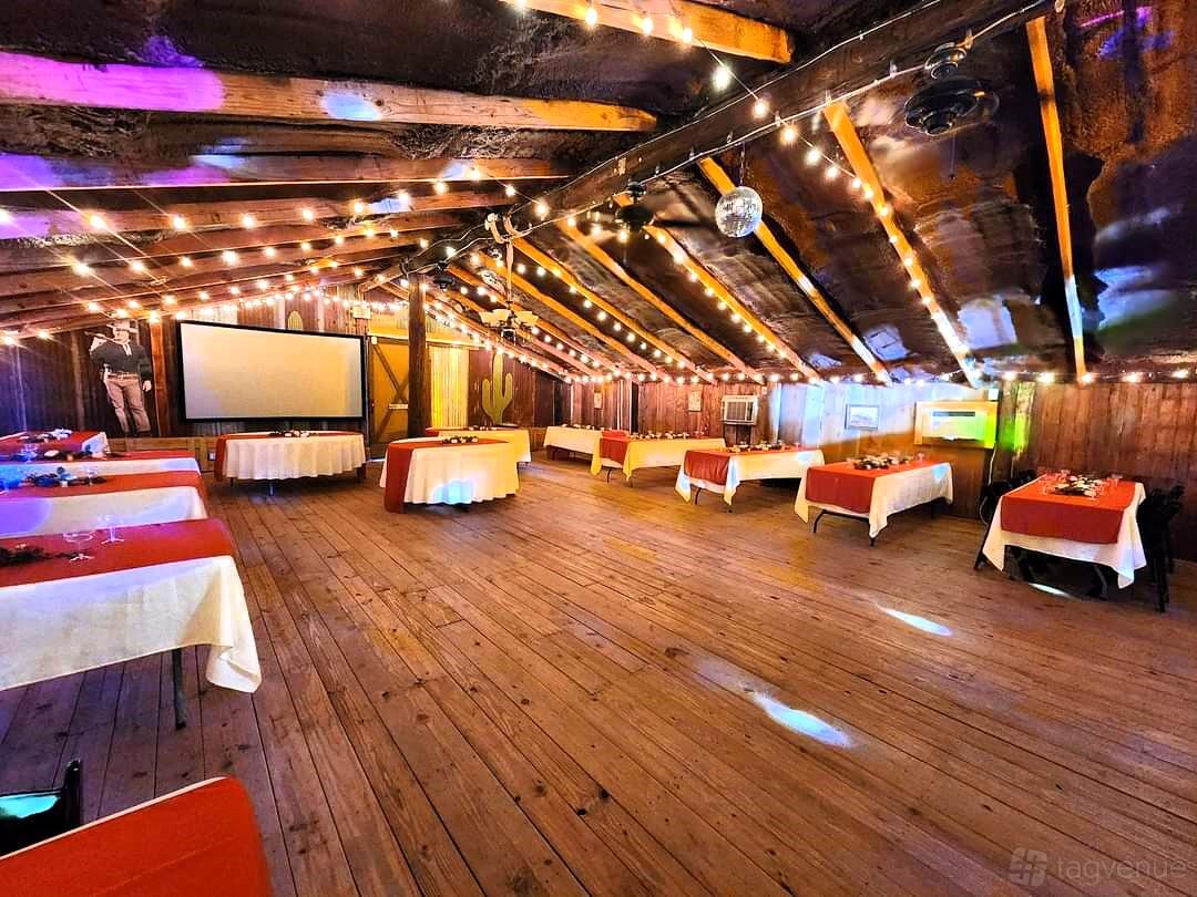 An event venue with exposed wooden beams, string lights, a disco ball, and round tables with red linens at Chisholm Trail Rustic Venue.