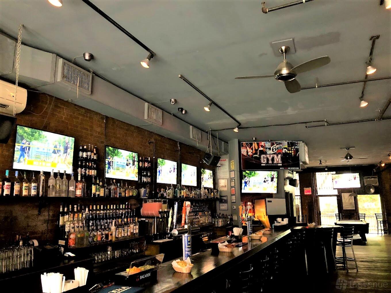 A bar with exposed brick walls, a long counter, multiple TVs, and overhead ceiling fans at Gym Sportsbar.