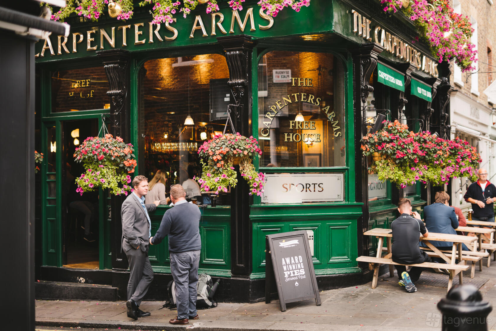 A traditional pub with green exterior, hanging flower baskets, and outdoor wooden benches at The Carpenters Arms.