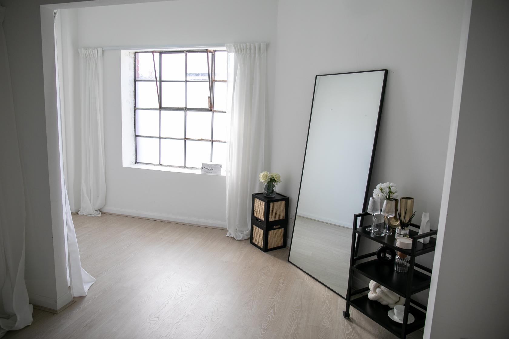 A photo studio with a large industrial window, white walls, a full-length mirror, and a shelving unit at Sugar Studio Bristol.