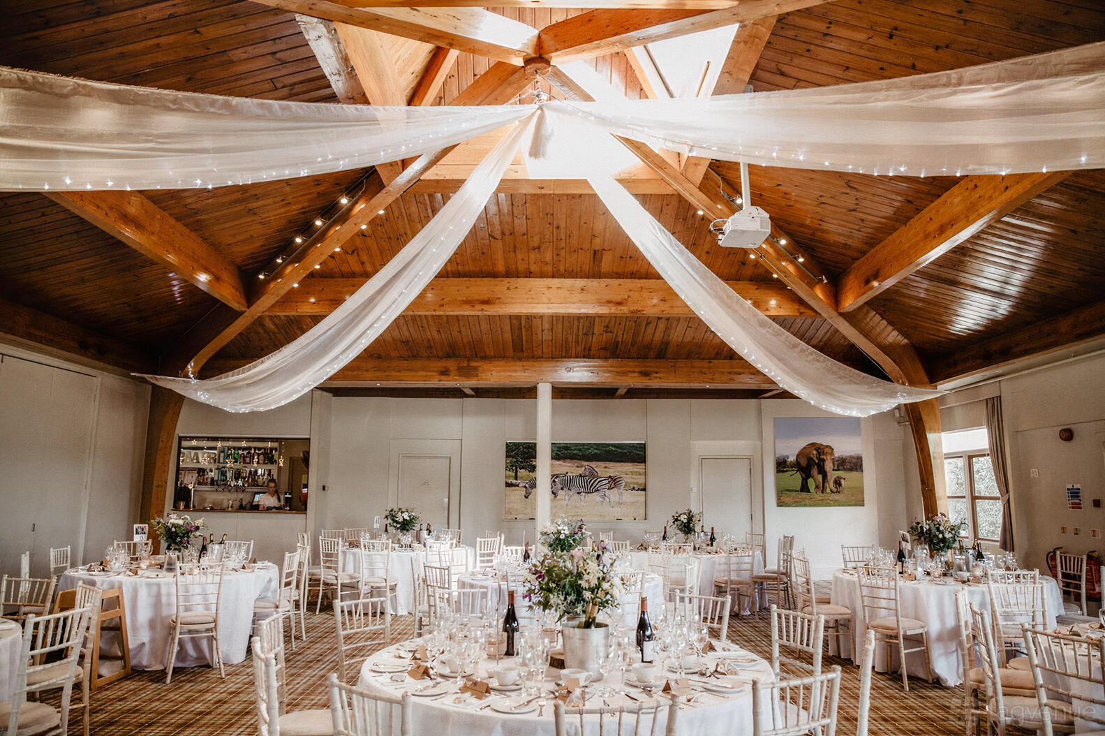 An event venue with draped fabric ceiling, wooden beams, and round tables set for dining at Woburn Safari Park Safari Lodge.