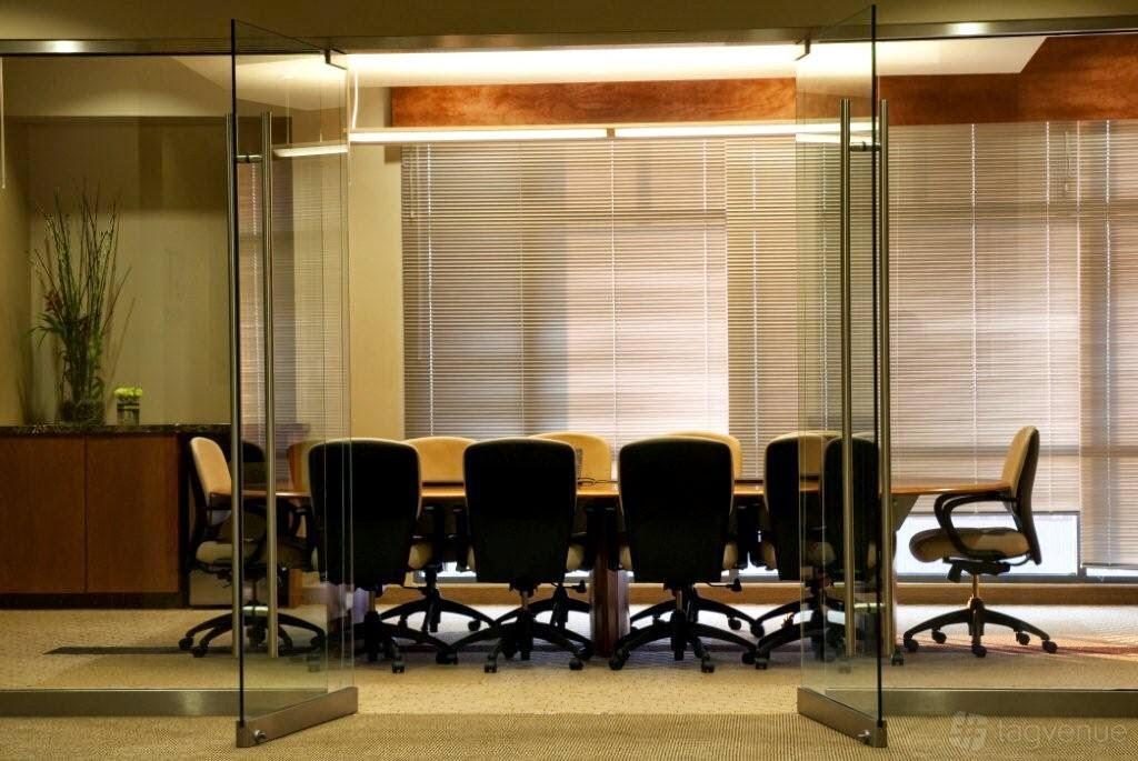 A meeting room with a long wooden table, ergonomic chairs, glass doors, and large covered windows at St Rose Executive Suites and Virtual Offices.
