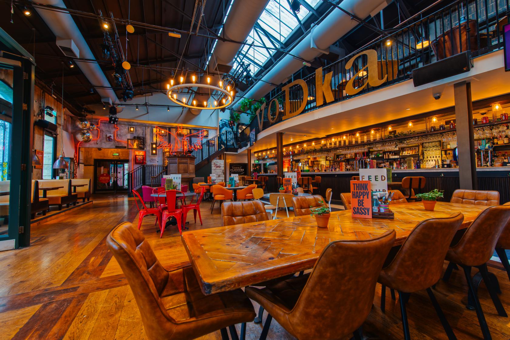 A bar with leather chairs, wooden tables, a mezzanine level, and a long backlit bar at Revolution Bristol.