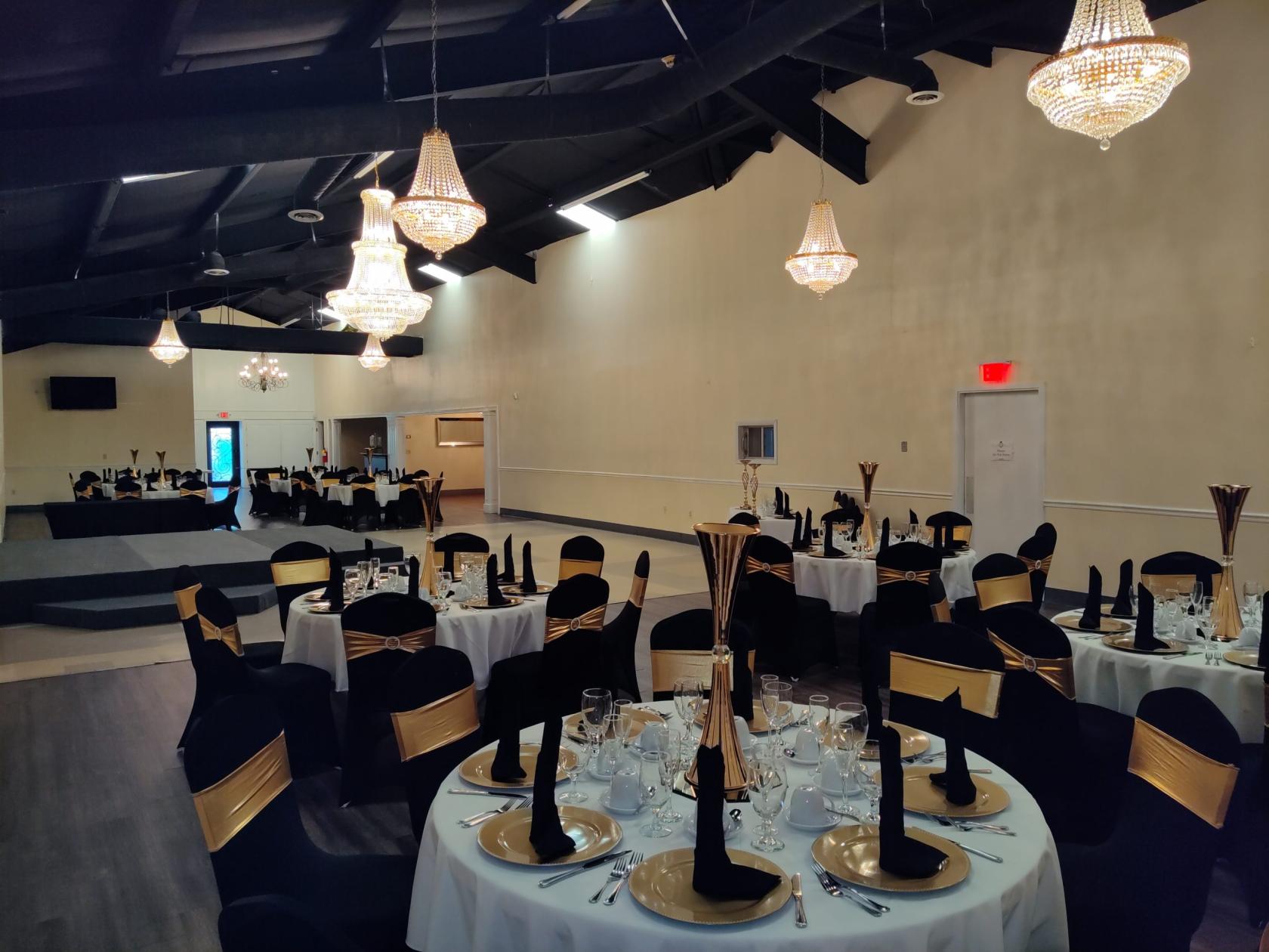 A ballroom with round tables dressed in black and gold linens, chandeliers, and a stage at JVC's Party Rentals & Event Hall.