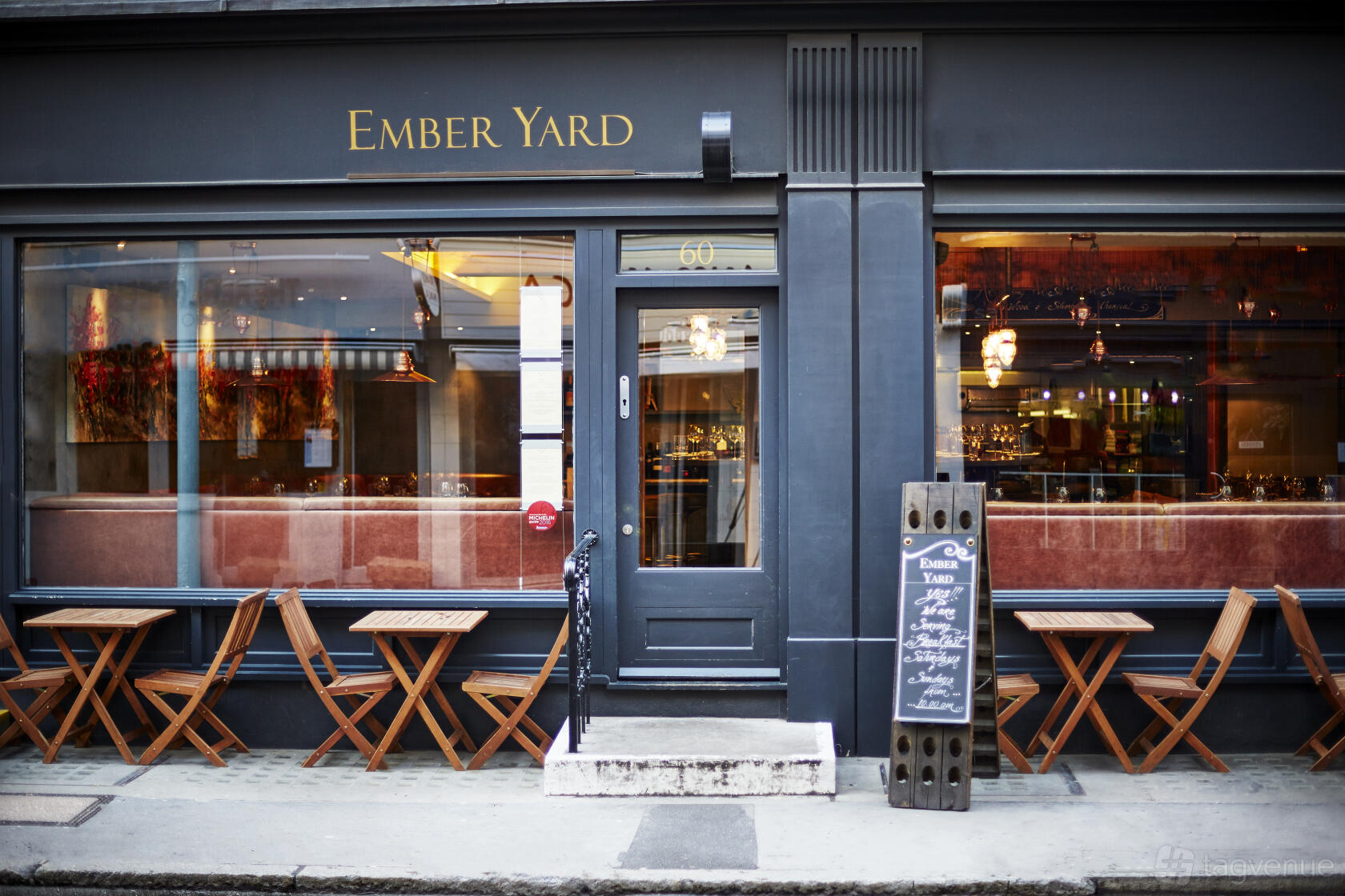 A restaurant exterior with large windows, black facade, and outdoor wooden tables at Ember Yard.