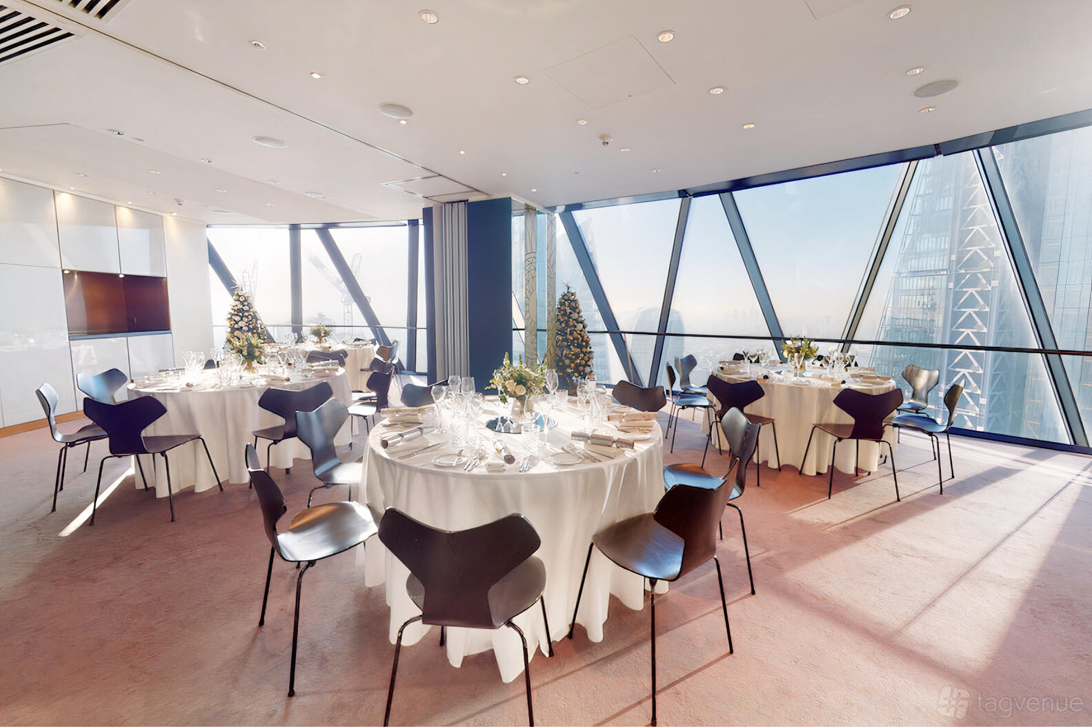 A function room with floor-to-ceiling glass walls, round tables with white linens, and city views at Searcys at The Gherkin.