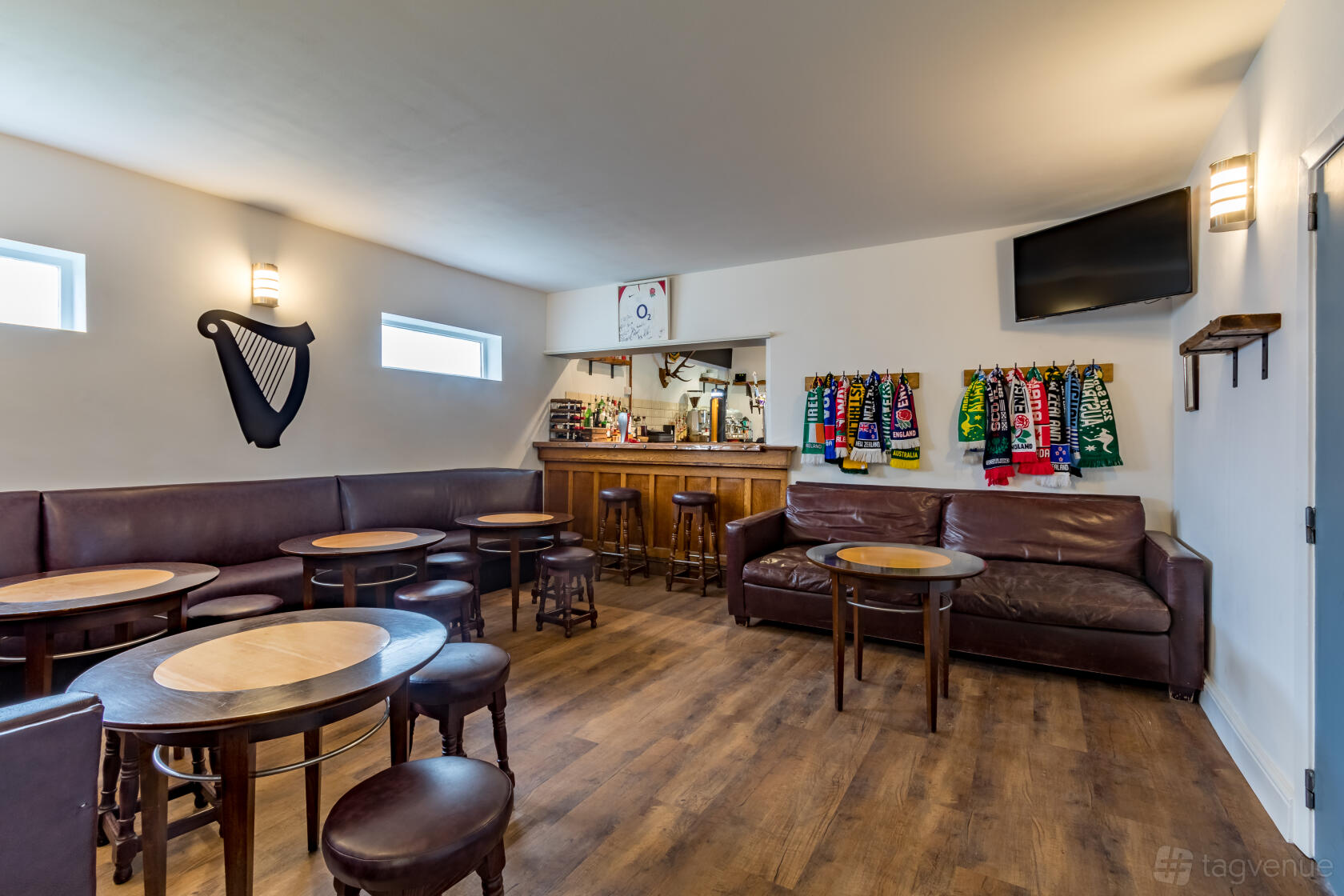A lounge with dark leather seating, round wooden tables, and sports scarves displayed behind the bar at WRFC BAR & COFFEE HOUSE.