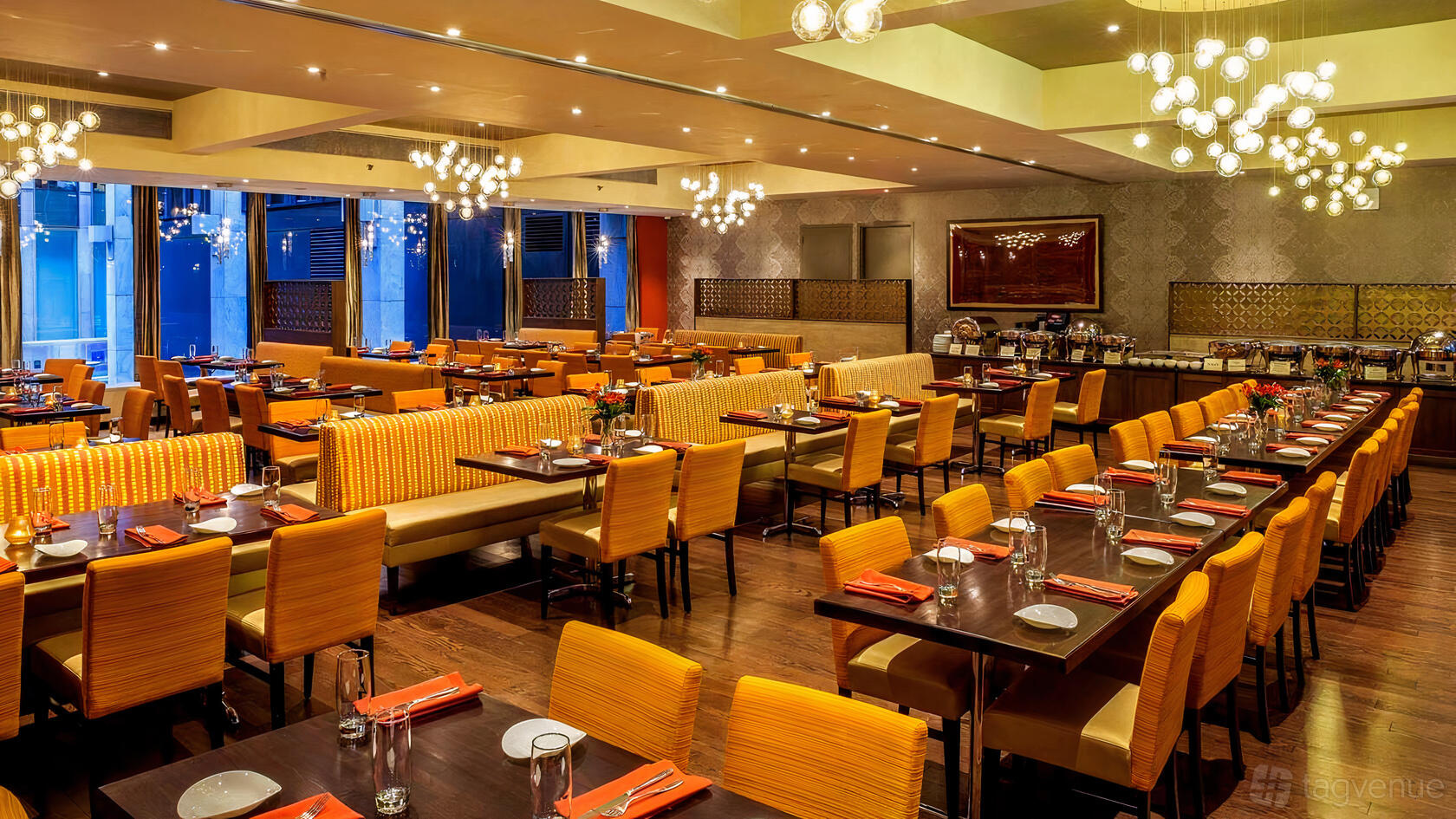 A restaurant dining room with orange upholstered chairs, booth seating, and modern chandeliers at Utsav.