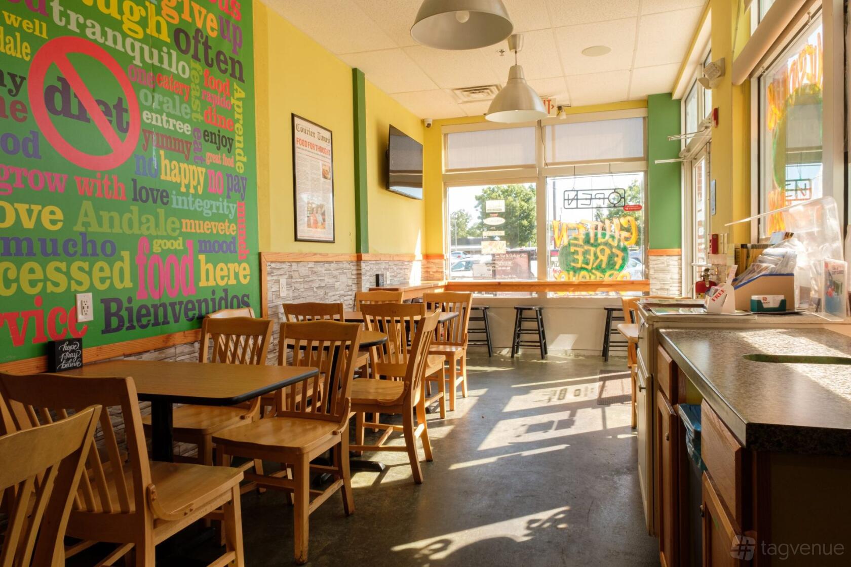 An event space with colorful wall art, wooden tables and chairs, and large sunlit windows at Andale Latino Grill.