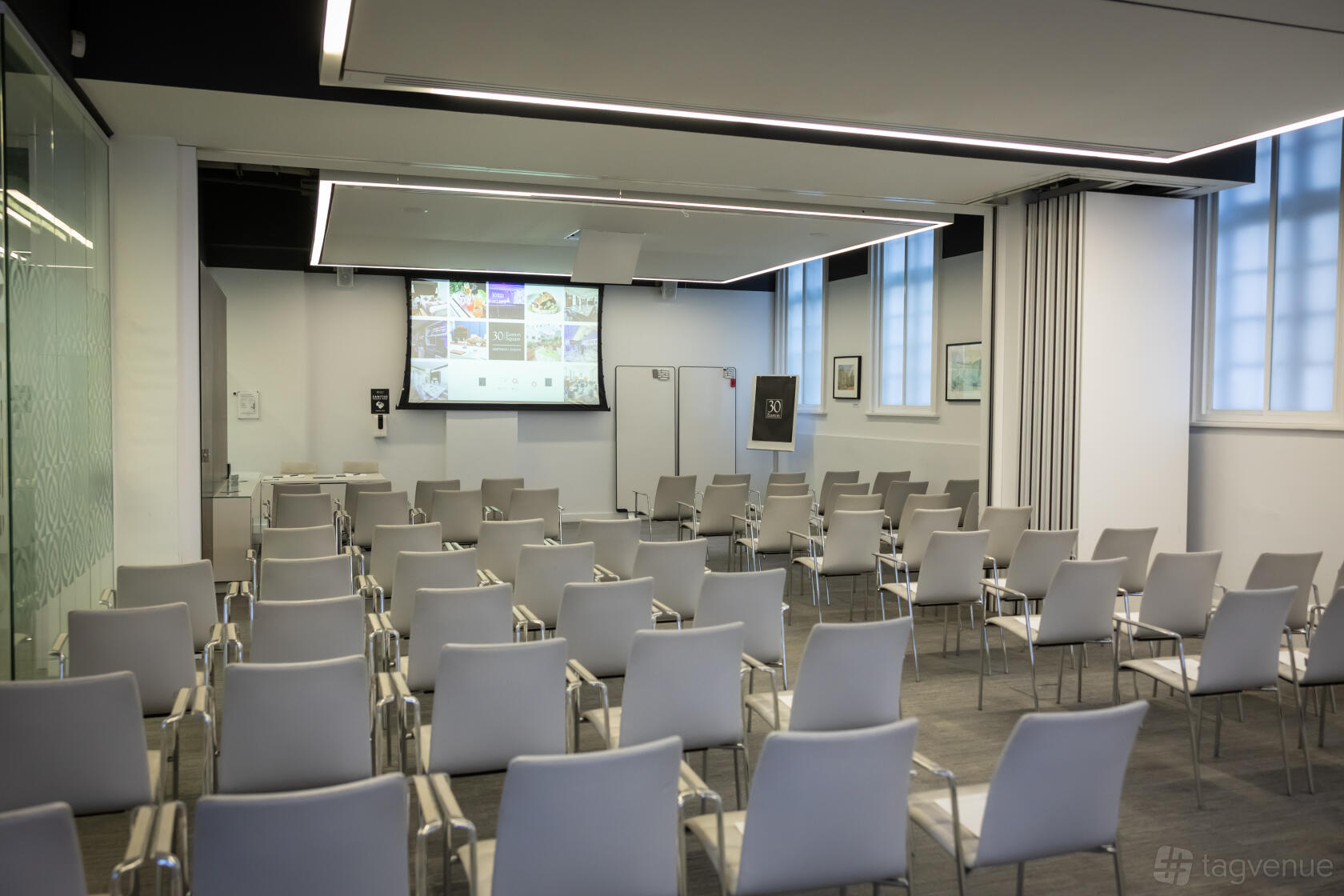 A meeting room with rows of grey chairs, ceiling-mounted projector, and large presentation screen at 30 Euston Square.