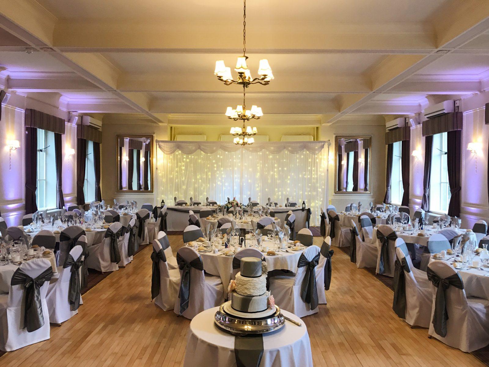 A country house dining room with round tables, white linens, draped chairs, and chandeliers at Hoyle Court.