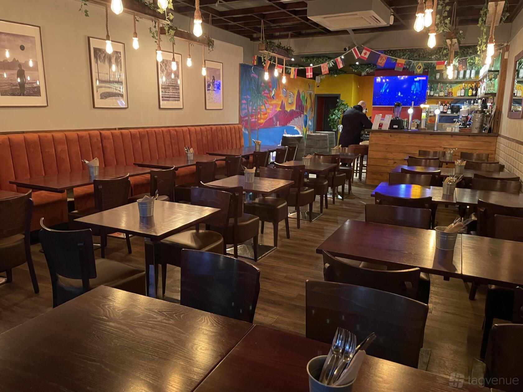 A restaurant with warm lighting, wood tables, orange banquette seating, and colorful wall art at Ipanema Restaurant and Bar.