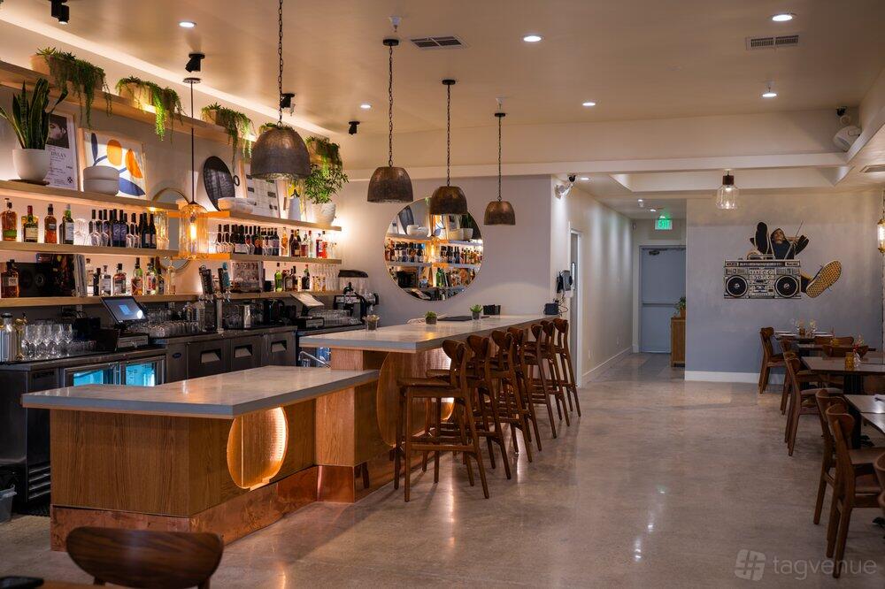 A restaurant with a long marble bar, wooden chairs, pendant lighting, and a decorated back wall at The Shop: Pizza and Cocktails.