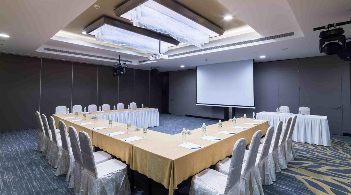 A function room with U-shaped tables covered in beige linens, bottled water, and a projector screen at The Chevrons.