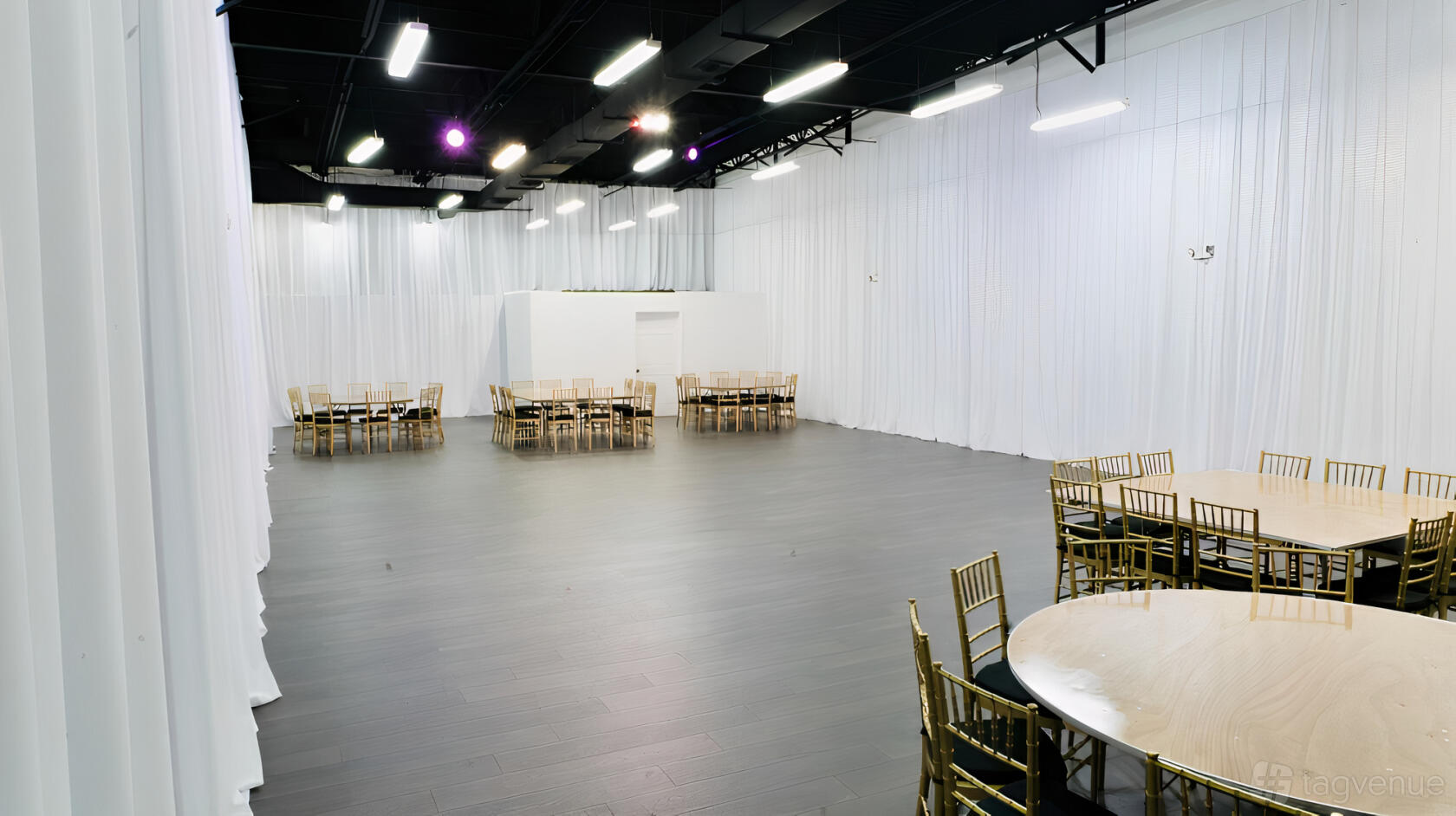 An event venue with white draped walls, dark wood floors, and round tables with gold chairs at Y-Squared Event Venue.