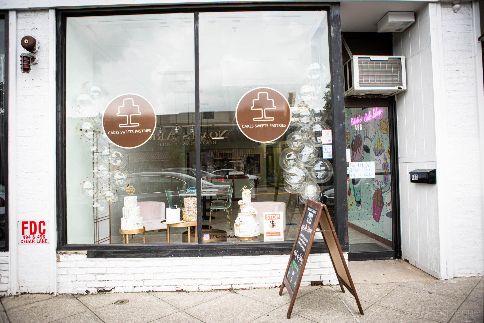 A café storefront with a large glass window, hanging cake shop signs, and decorated cakes on display at Krisota's Cake Shop.