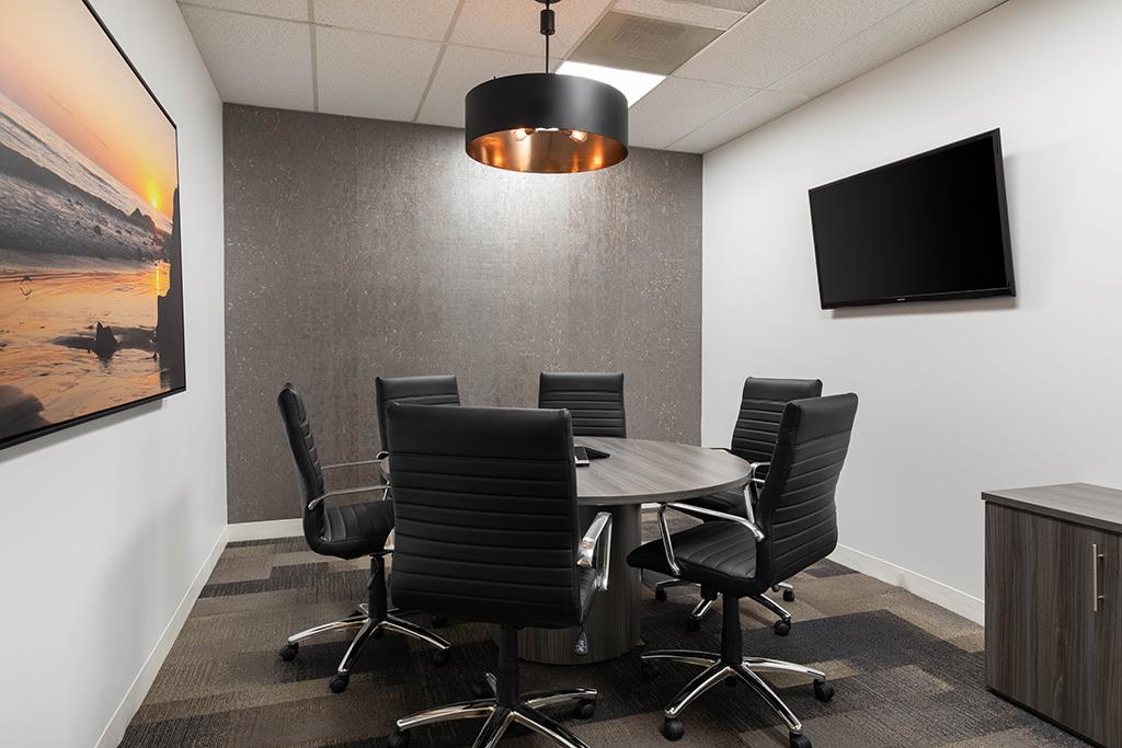 A boardroom with a round table, black leather chairs, wall-mounted TV, and pendant lighting at Premier Workspaces - El Segundo.