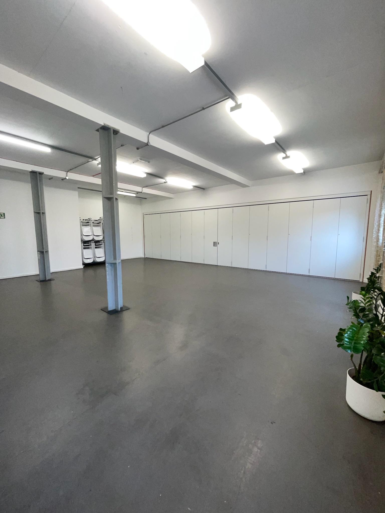A function room with polished concrete floors, white walls, ceiling strip lights, and stacked folding chairs at Boathouse Studios, Barking.