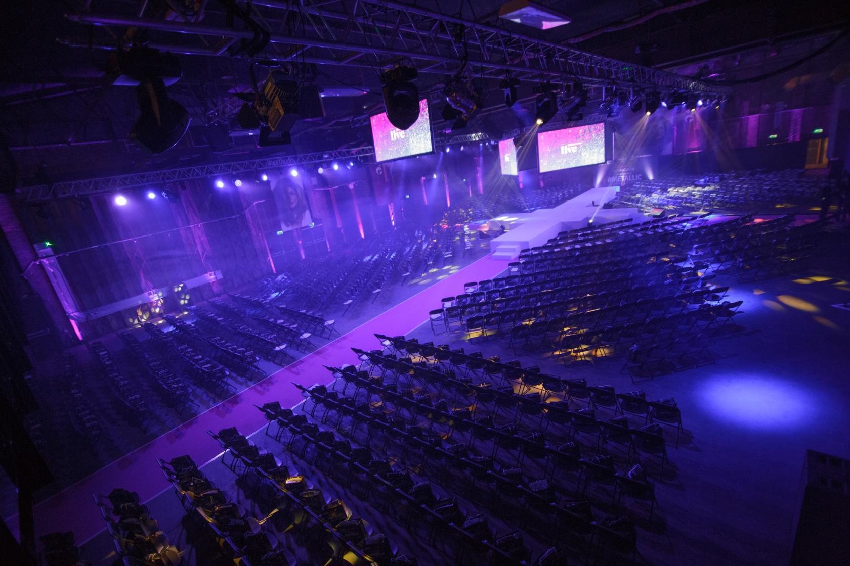 A warehouse with rows of black chairs facing a stage and purple lighting at Victoria Warehouse.