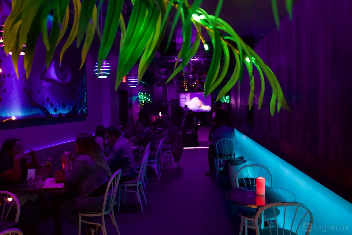 A dimly lit bar with neon purple and blue lighting, wall art, and tables with teal chairs at Doha Bar Lounge.