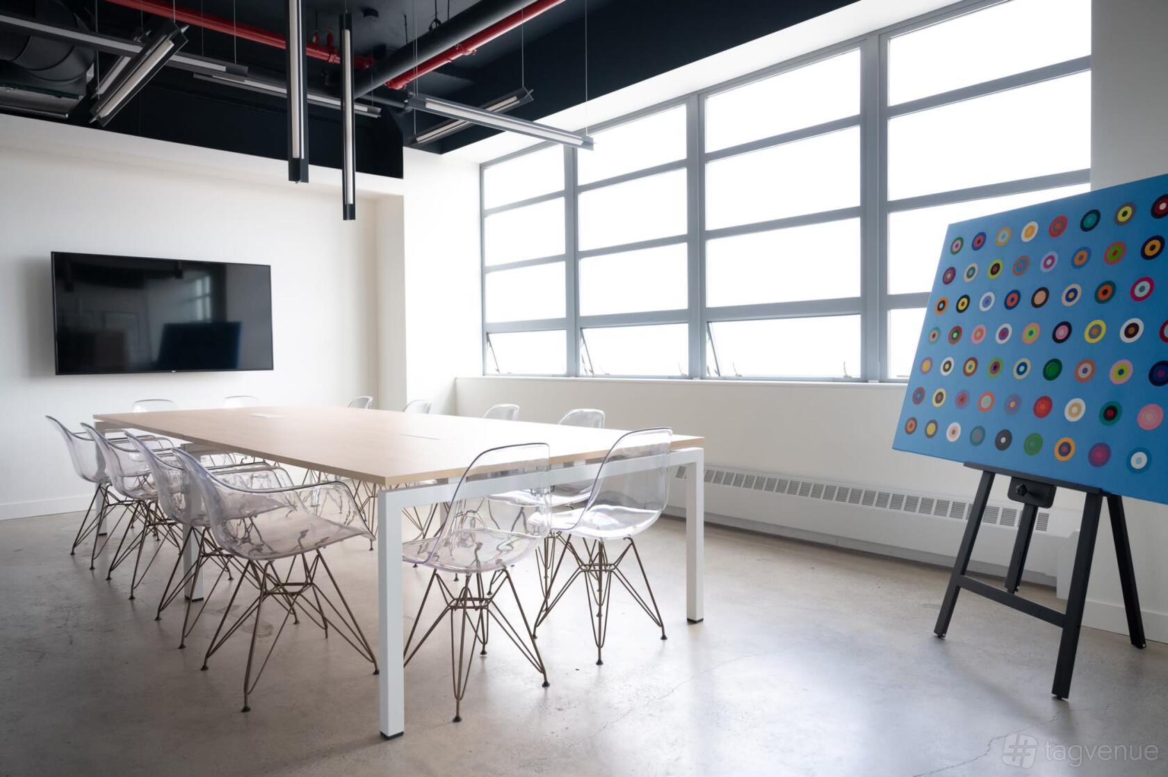 A conference room with clear chairs, a long table, large windows, a wall-mounted screen, and an abstract painting at Private Loft Gallery.