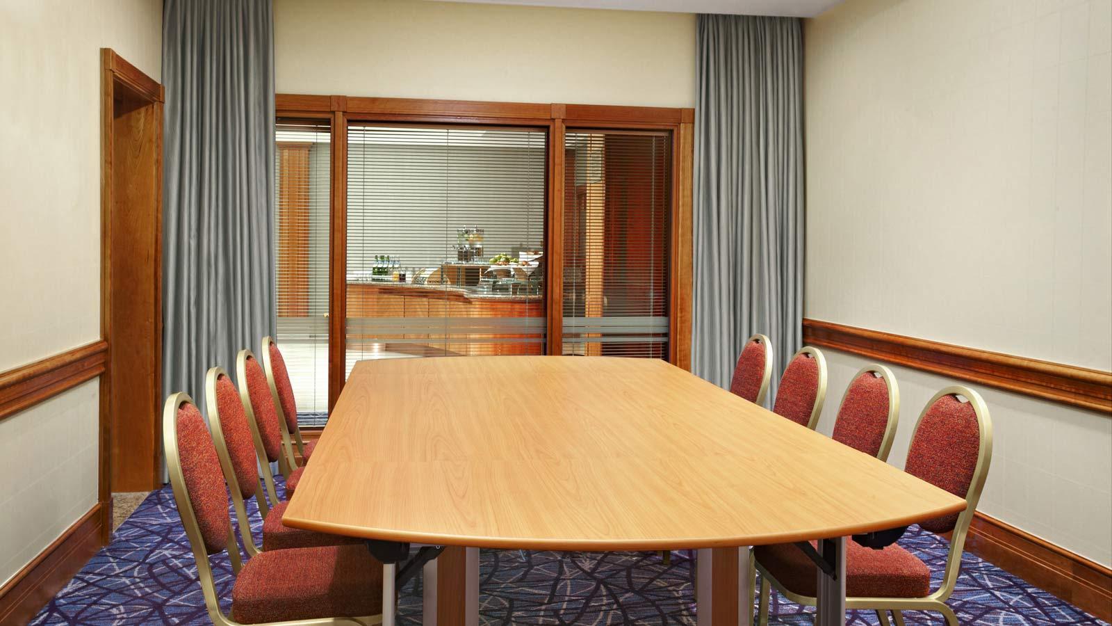A meeting room with a long wooden table, red cushioned chairs, carpeted floor, and window blinds at Sheraton Skyline Hotel London Heathrow.
