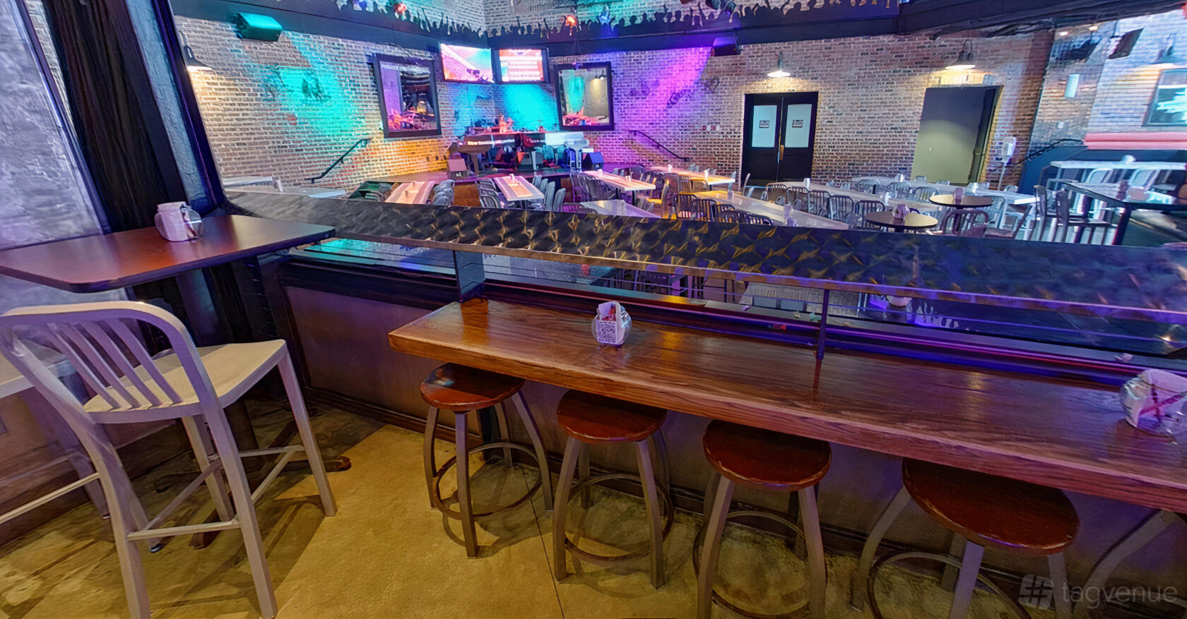 A bar with a long wooden rail, metal stools, exposed brick walls, and colorful stage lighting at Bobby McKey's Dueling Piano Bar.