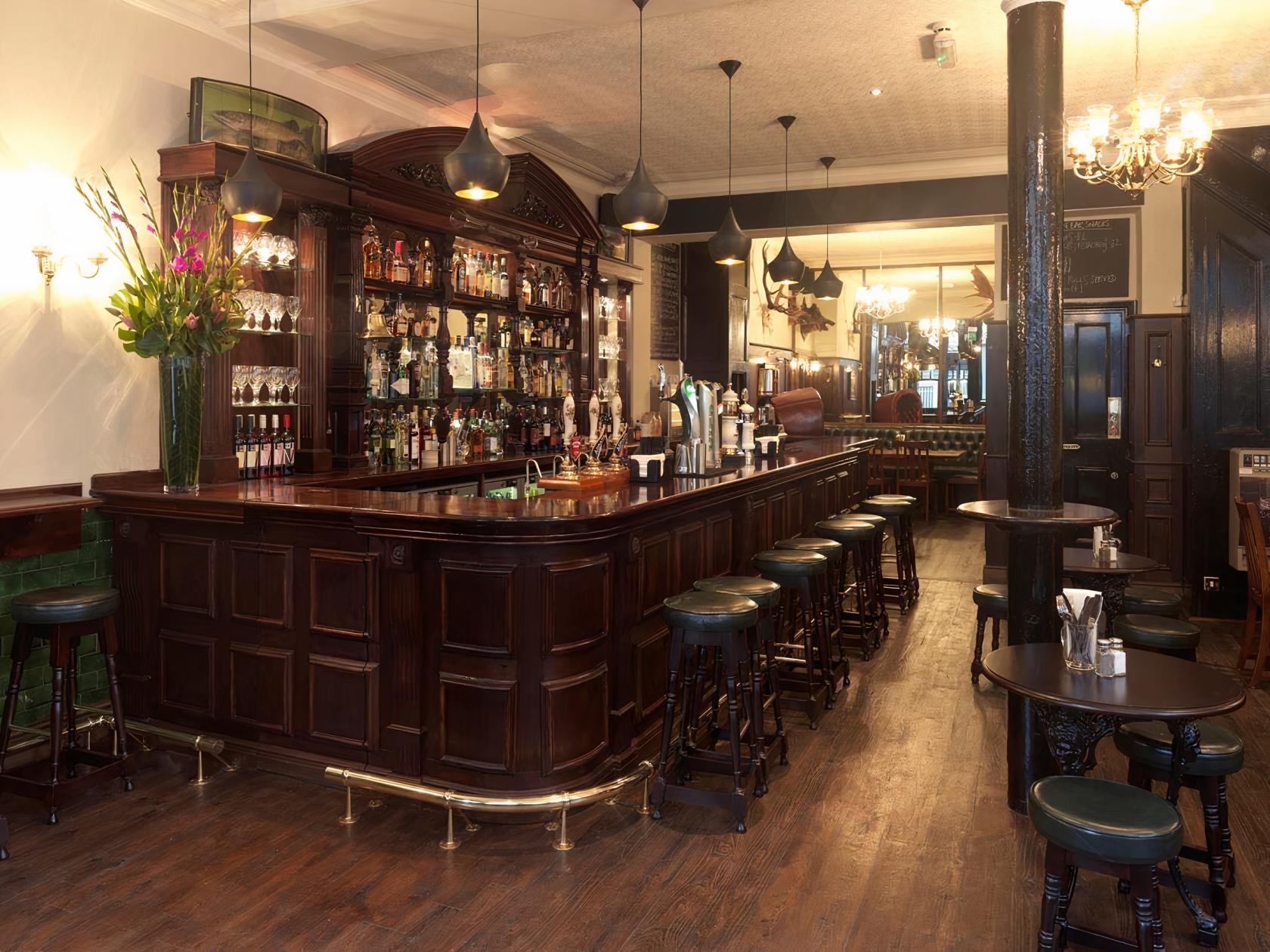 A traditional pub with a dark wood bar, vintage bar stools, and mirrored back shelves at The Hat and Tun.