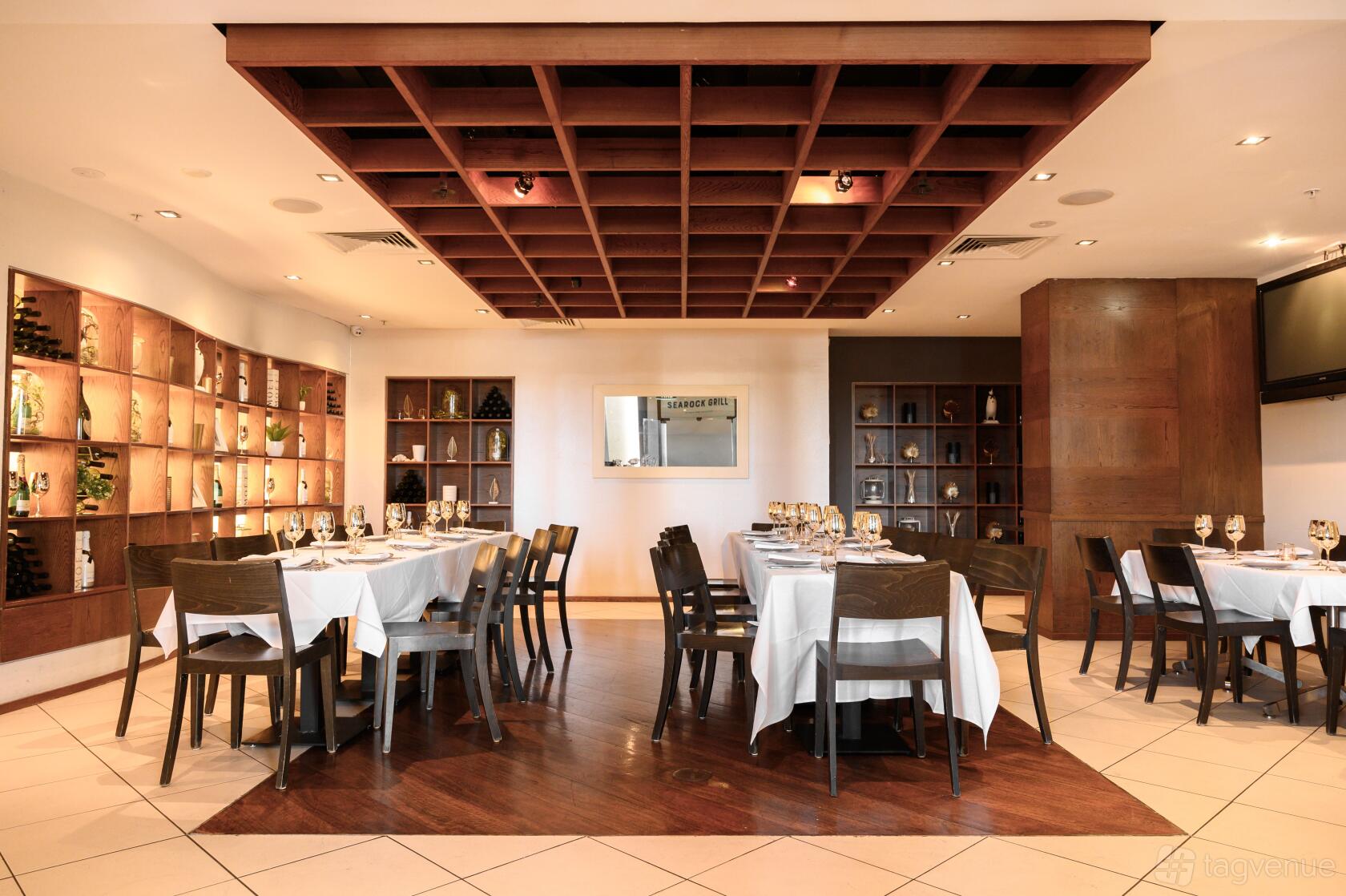 A restaurant function room with wine bottle display shelves, square wooden tables, and white tablecloths at Searock Grill.