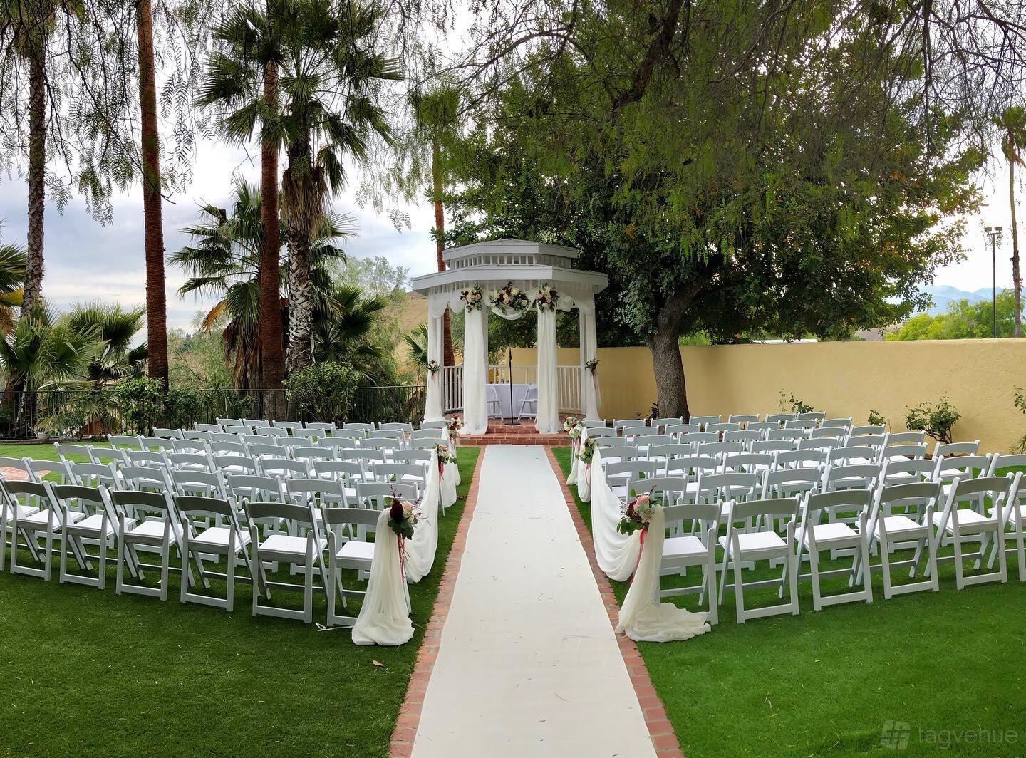 An outdoor event venue with a white gazebo, rows of white chairs, floral arrangements, and a paved aisle at Hilltop Collection San Bernardino.