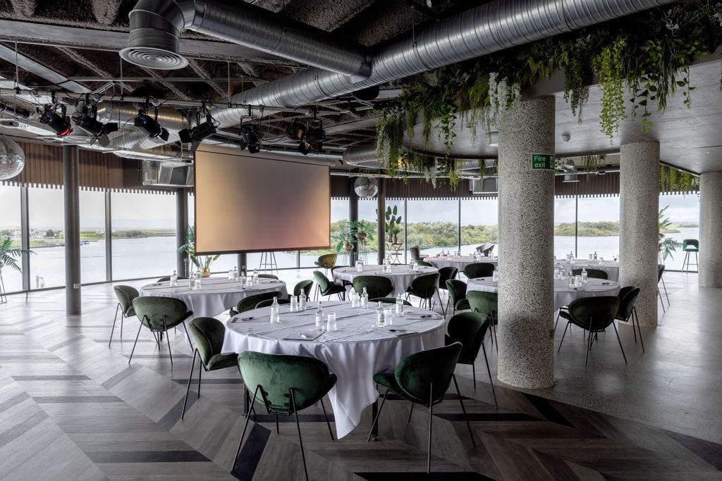 An event space with round tables, green velvet chairs, floor-to-ceiling windows, and water views at Waterfront Southport Hotel.
