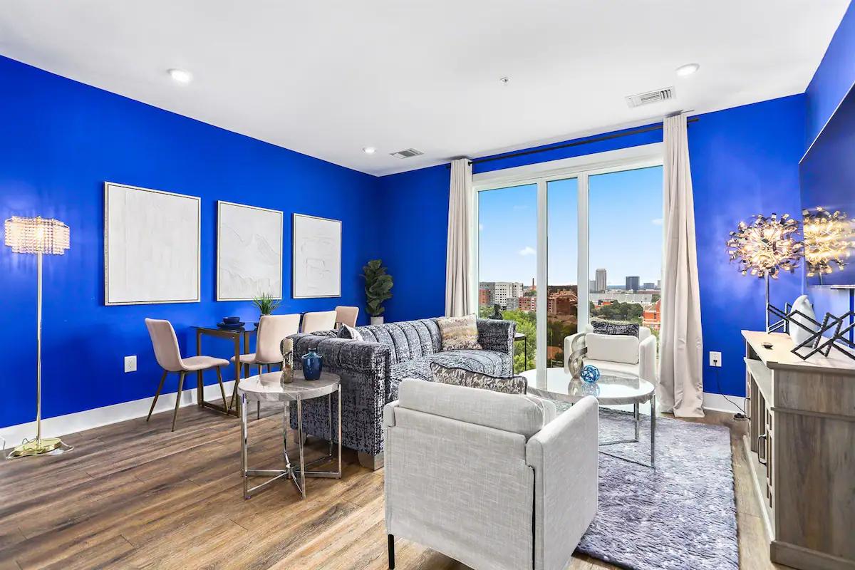 An apartment living area with bold blue walls, large windows, city views, modern seating, and wood floors at 'Midnight Oasis' Apartment.