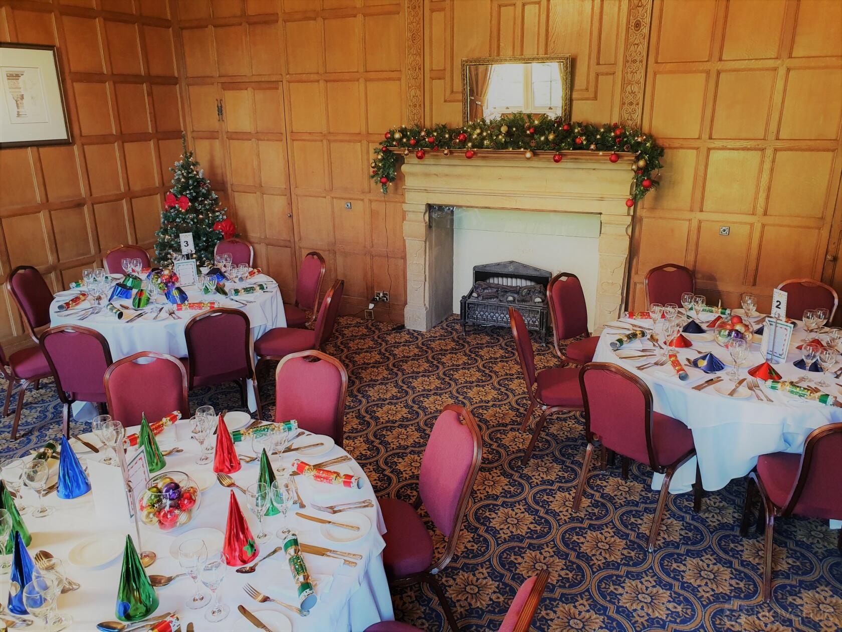 A hotel event room with wood panelled walls, a decorated fireplace, and round tables set for dining at Dumbleton Hall Hotel.