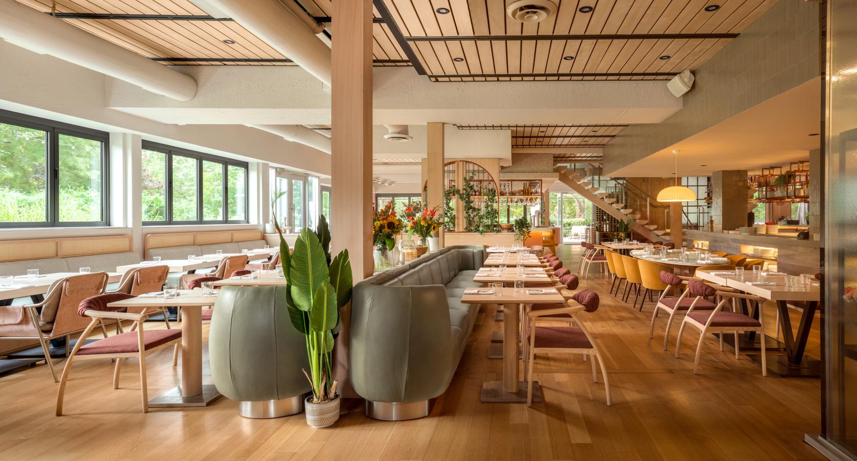 A restaurant with floor-to-ceiling windows, wood panel ceilings, green banquette seating, and round tables at Toronto Beach Club.