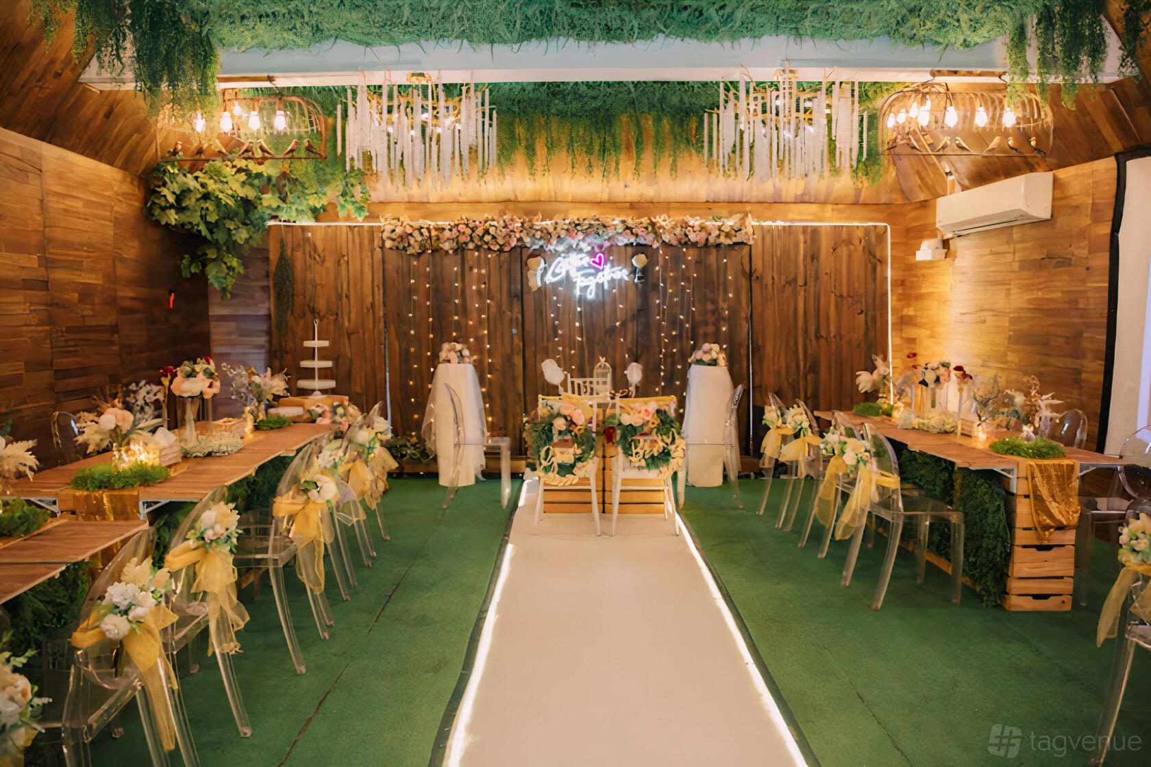 A hall with a green carpeted aisle, wooden walls, hanging greenery, and floral table decorations at Into The Woods.