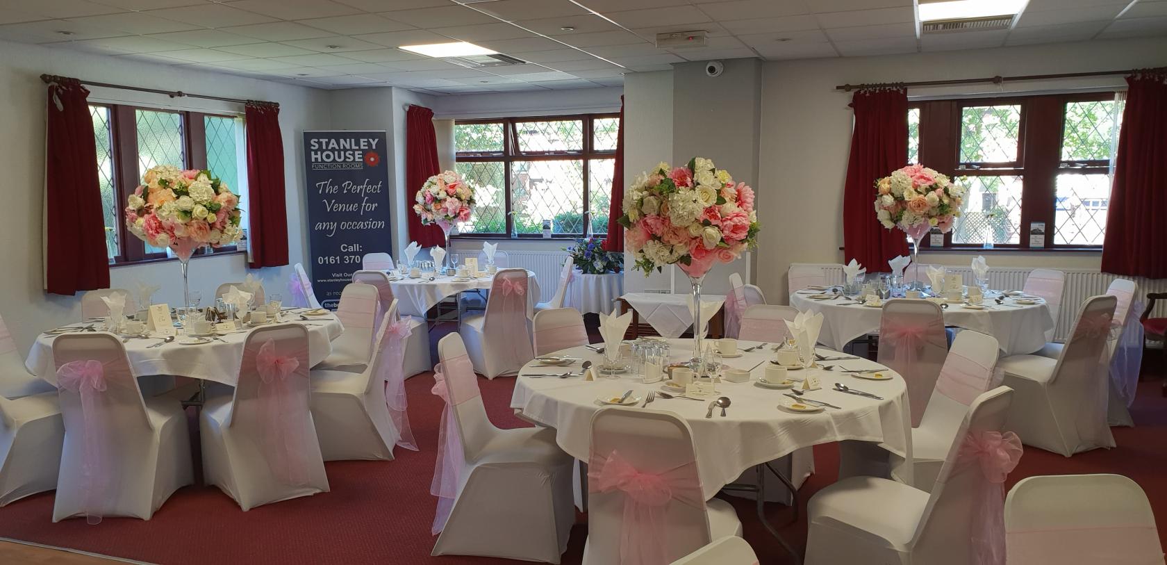 A function room with round tables, white chair covers with pink sashes, and floral centerpieces at Stanley House Function Rooms.