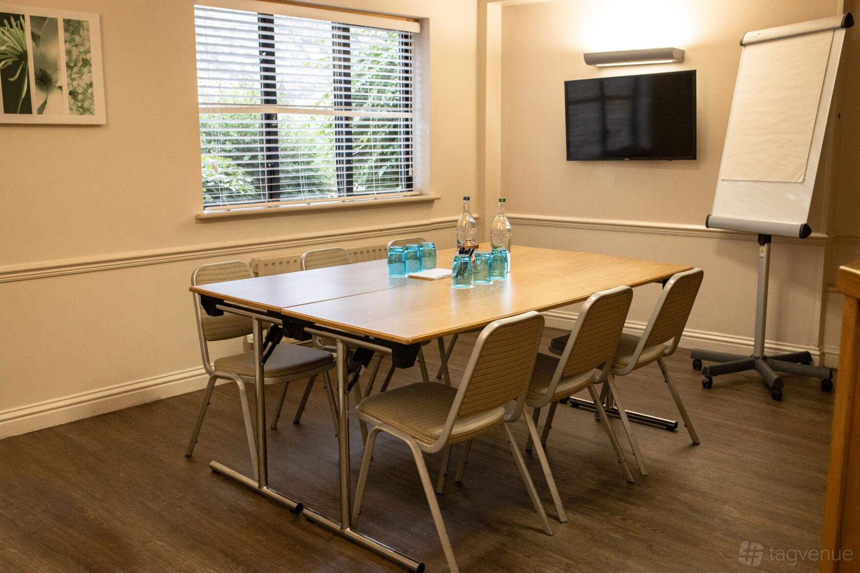 A meeting room with a rectangular table, six chairs, flipchart, wall-mounted TV, and natural light at Voco Oxford Thames.