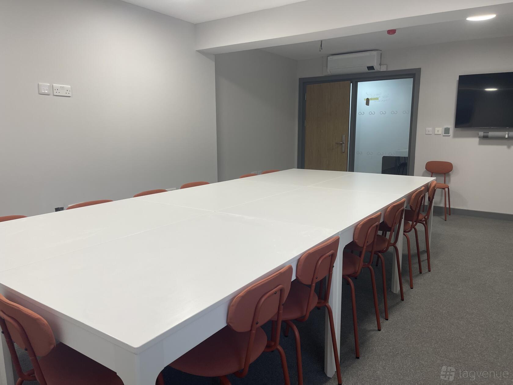A coworking boardroom with a long white conference table, red chairs, and a wall-mounted screen at Collaborative One - Flexible Spaces.