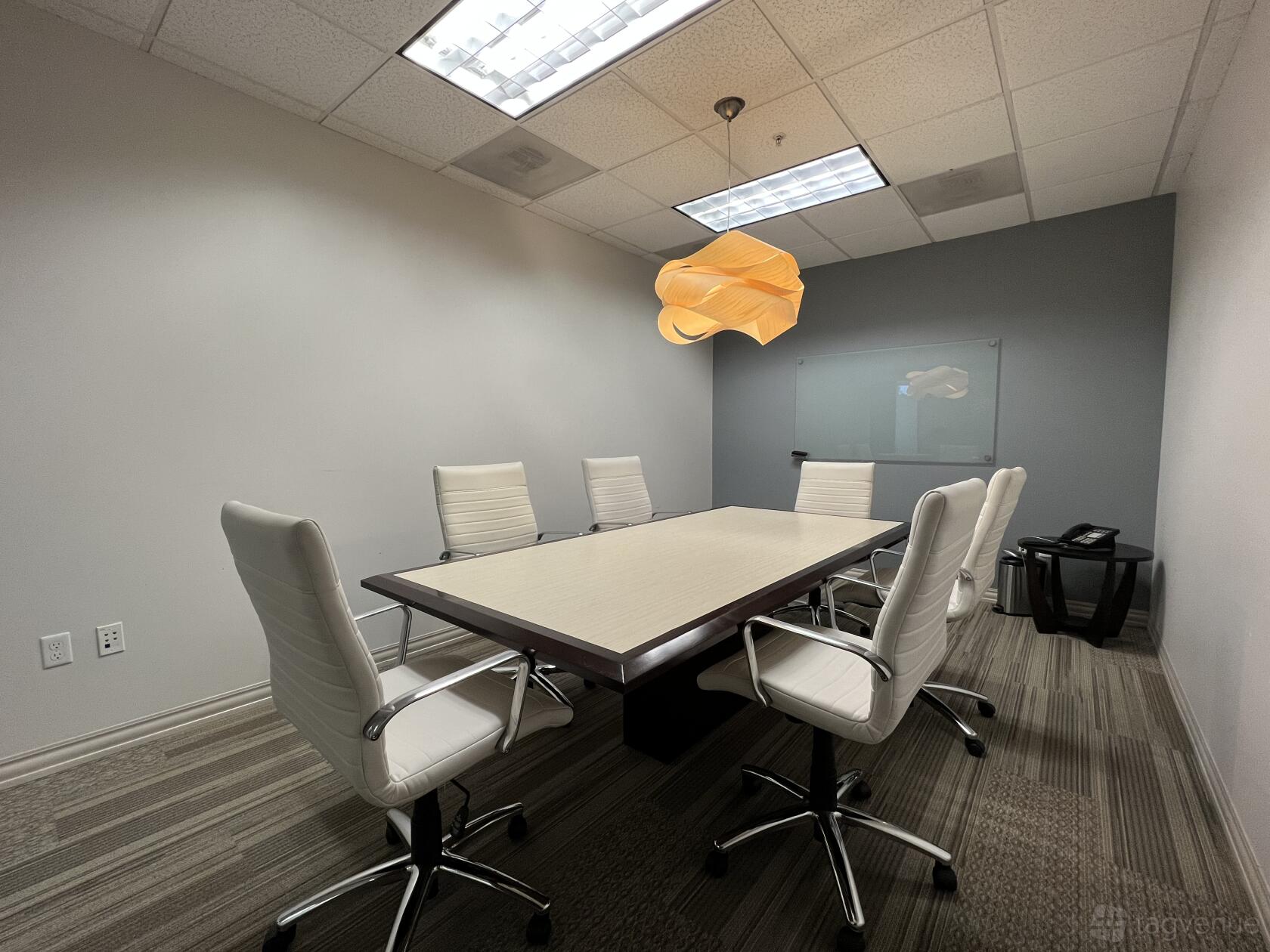 A meeting room with a rectangular conference table, six white chairs, a glass whiteboard, and pendant lighting at Premier Workspaces - Foothill Ranch.