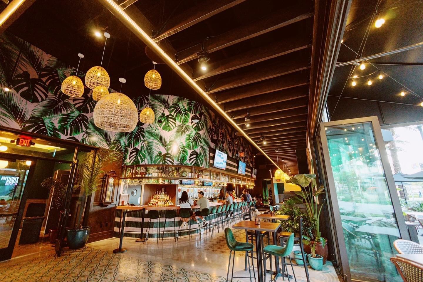 A bar in a theatre with tropical wallpaper, rattan pendant lights, and an open ceiling at Dania Improv.