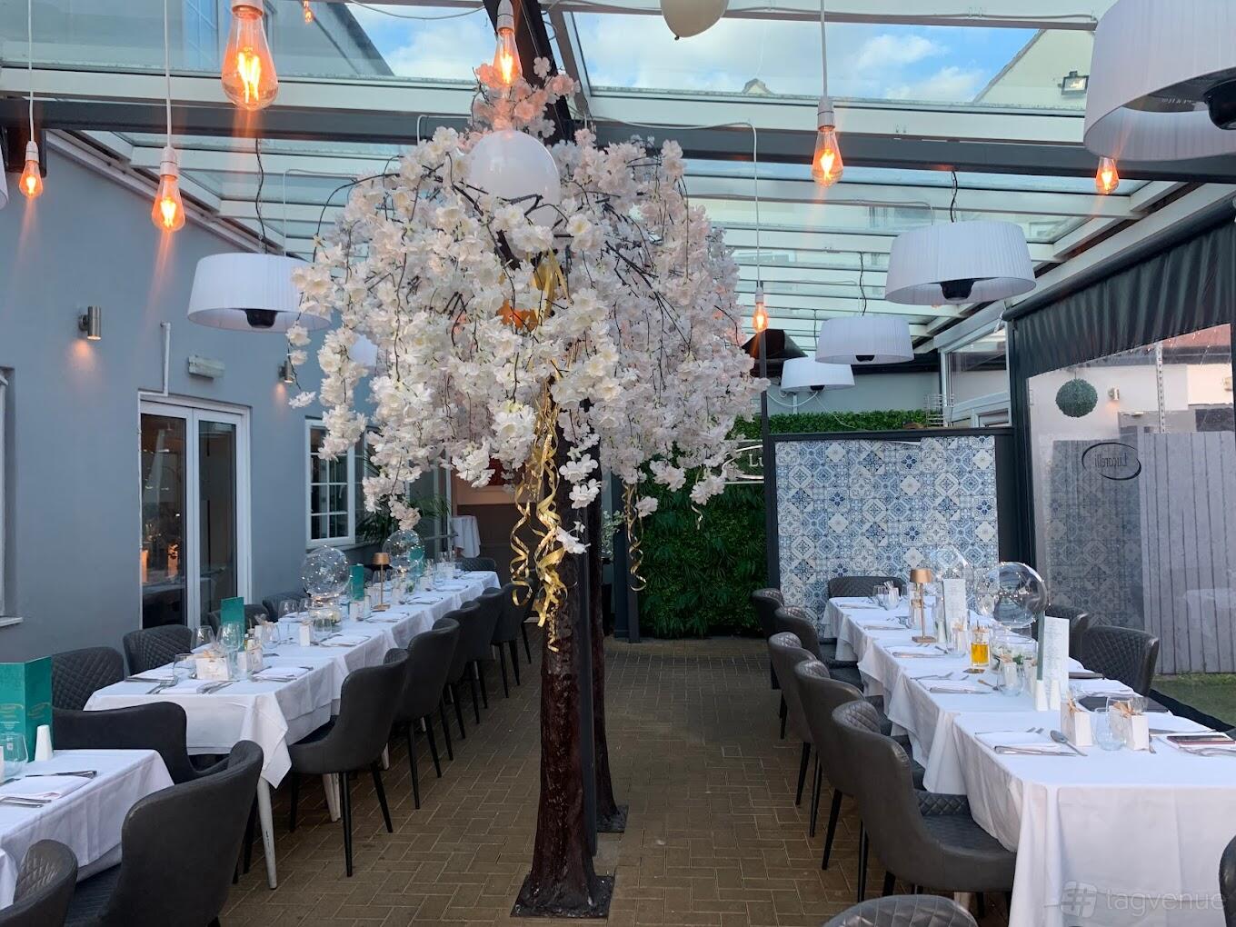 A dining room with a glass ceiling, long tables with white linens, and a decorative blossom tree at Lucarelli West Bromwich.