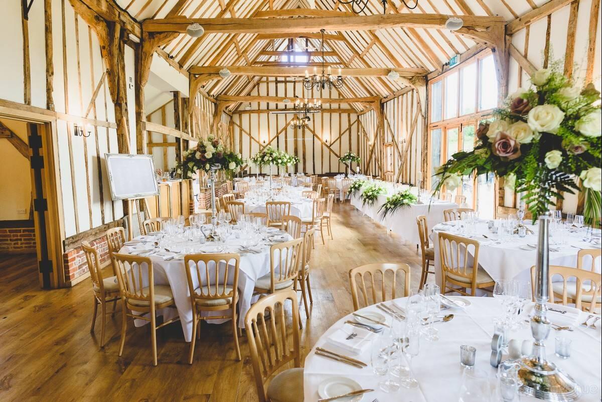 An event space with exposed wooden beams, tall windows, and round tables with floral centerpieces at Bruisyard Country Estate.