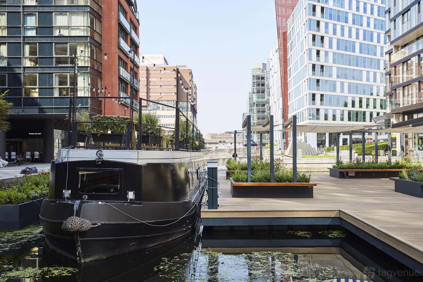 A hotel boat moored on a canal with a wood deck terrace and modern buildings at The Boathouse London - Paddington West.