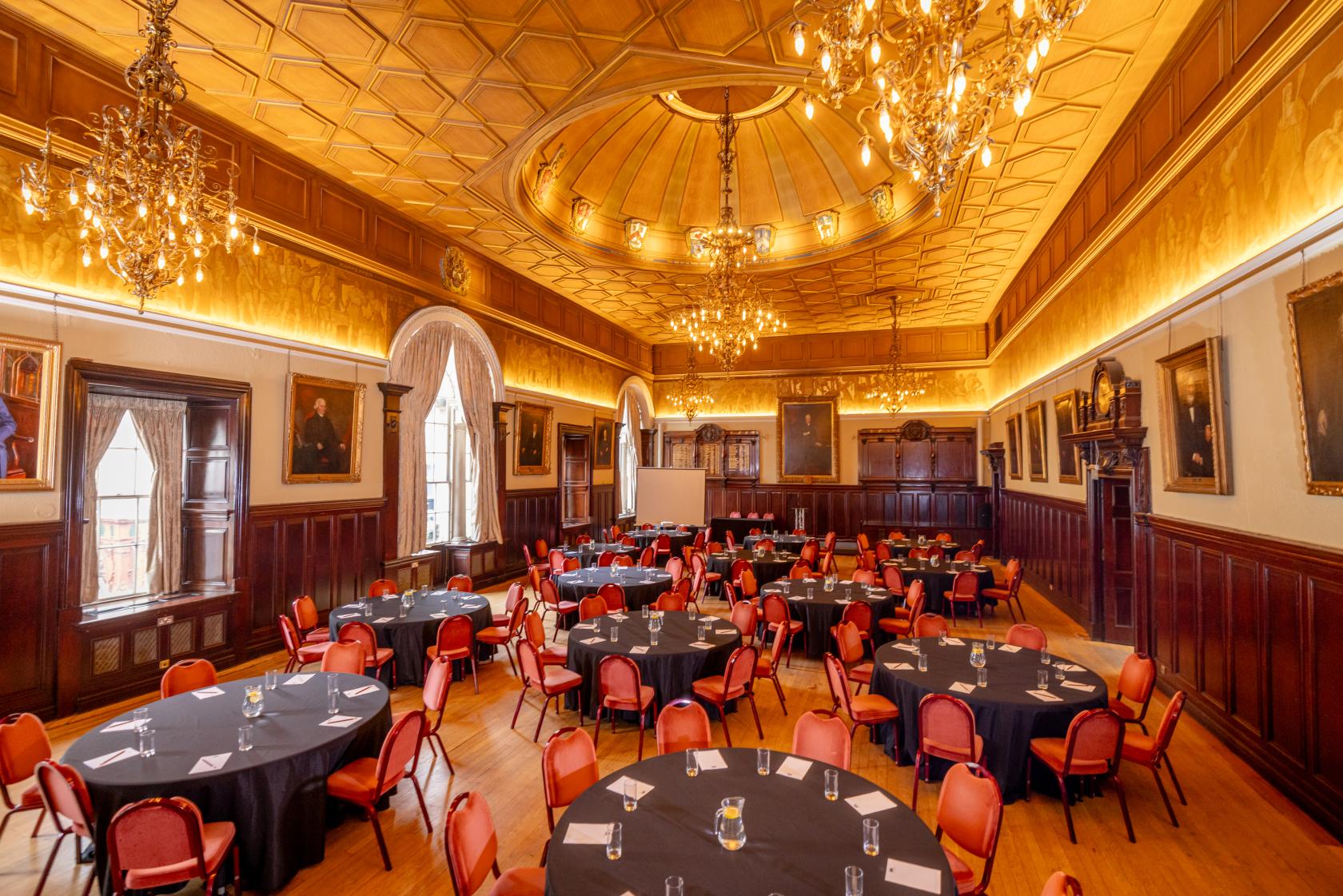 A grand hall with gilded ceilings, chandeliers, large arched windows, and round tables at Trades Hall of Glasgow.