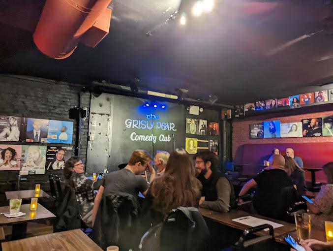 A bar with black brick walls, comedy club signage, and wall art with closely arranged tables at The Grisly Pear Greenwich Village.